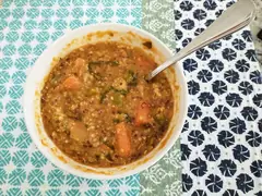 Бяла купа с гъста супа от леща и киноа с парчета морков и зеленчуци, с метална лъжица